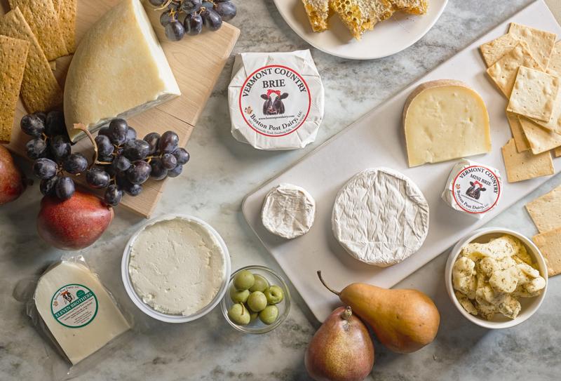 An overhead view of an artisanal cheese platter spread across a marble surface, featuring various wheels and wedges of cheese, crackers, purple grapes, green olives, pears, honeycomb, and wrapped packages of Vermont Country Brie.