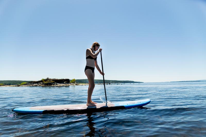 A person in a black bikini stands on a blue paddleboard, using a long paddle to steer through calm, clear water on a bright, sunny day, with a small rocky island visible in the background.