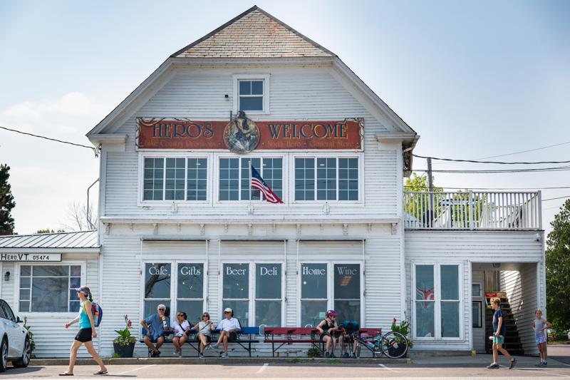 The front exterior of Hero's Welcome, a large white two-story country store with signs for a cafe, gifts, beer, deli, and wine, where pedestrians walk past and patrons sit on benches outside.