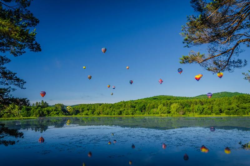 Colorful hot air balloons float in a clear blue sky over a calm lake and forested hills, reflecting on the water.