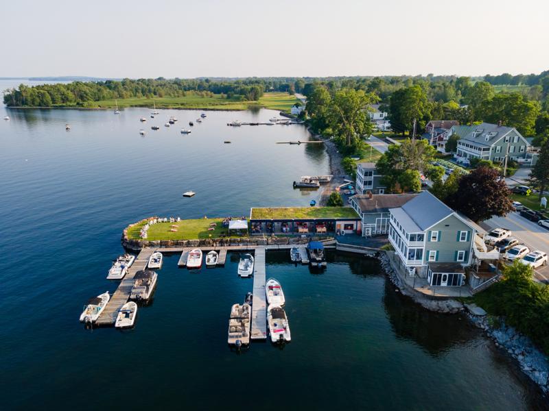 An aerial view of a lakeside marina and country store with a wooden boat dock filled with small motorized boats and pontoon boats, bordered by a green lawn and a two-story building alongside a calm bay.