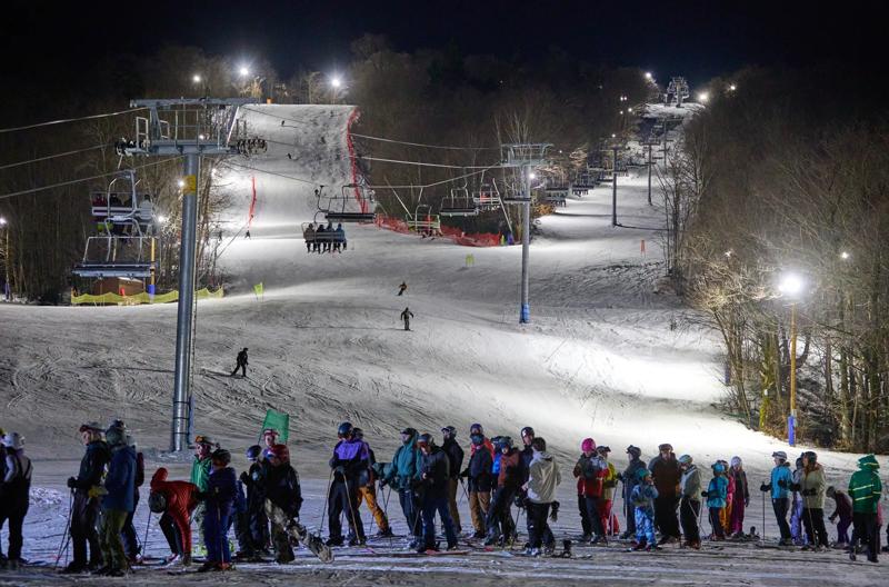 A crowd of skiers and snowboarders gathering at night at the base of a brightly illuminated ski slope with operational chairlifts.