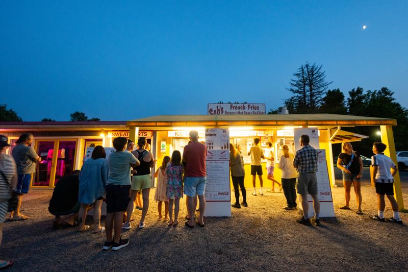 A crowd of people stands in lines at dusk outside a brightly lit outdoor food stand with signs for French fries, burgers, and ice cream under a deep blue twilight sky.