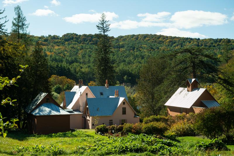 An elevated view of a rural homestead featuring light-colored buildings with metal roofs nestled in a valley, surrounded by dense evergreen trees and rolling green hills under a partly cloudy sky.