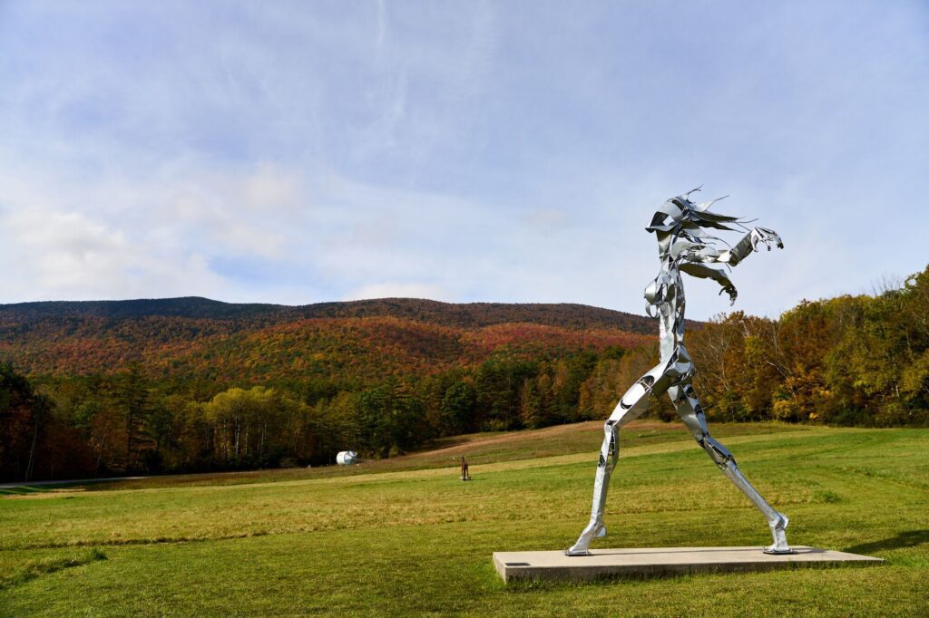 In the distance, a person walks alongside a sculpture of a much larger person running, seen outside in the fall.