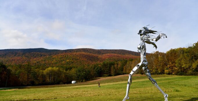 In the distance, a person walks alongside a sculpture of a much larger person running, seen outside in the fall.