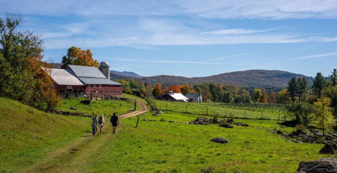 Family walks along a dirt trail towards a red barn with a silo. The grass is green and the surrounding trees are bright with red, orange, and yellow leaves.