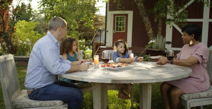 A family of four sits around an circular dining table outside. Appetizers and wine glasses sit on the table while the parent passes out plates.