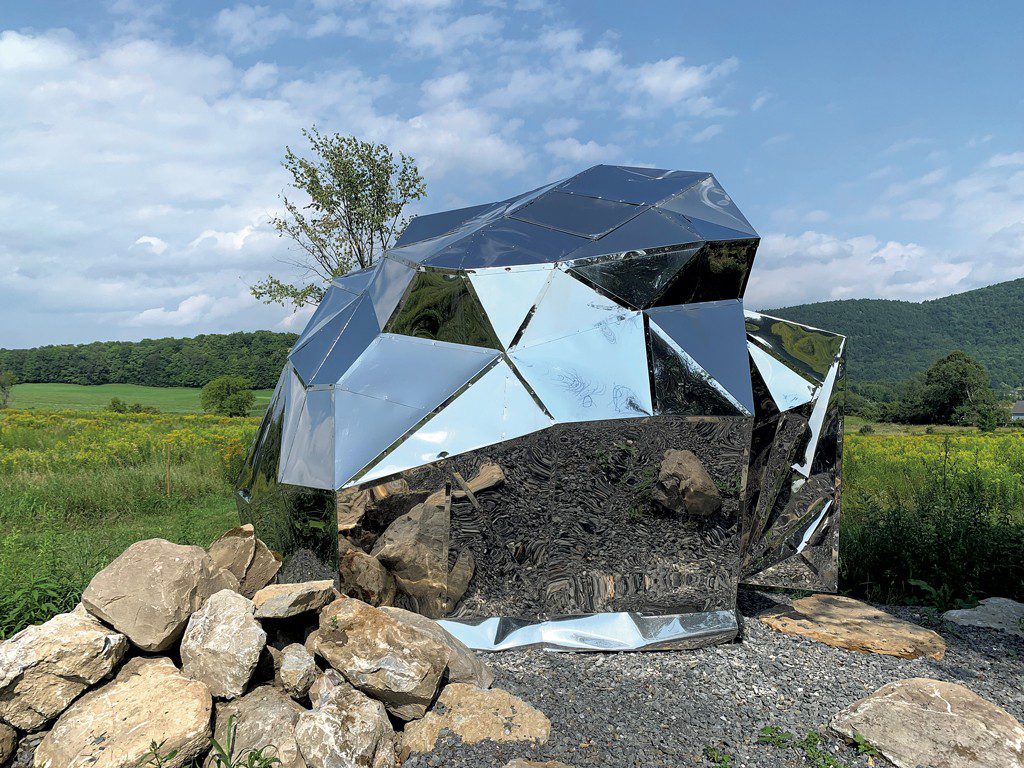A large, geometric mirrored sculpture sitting outdoors in a grassy field, reflecting the surrounding sky, ground, and distant green hills.