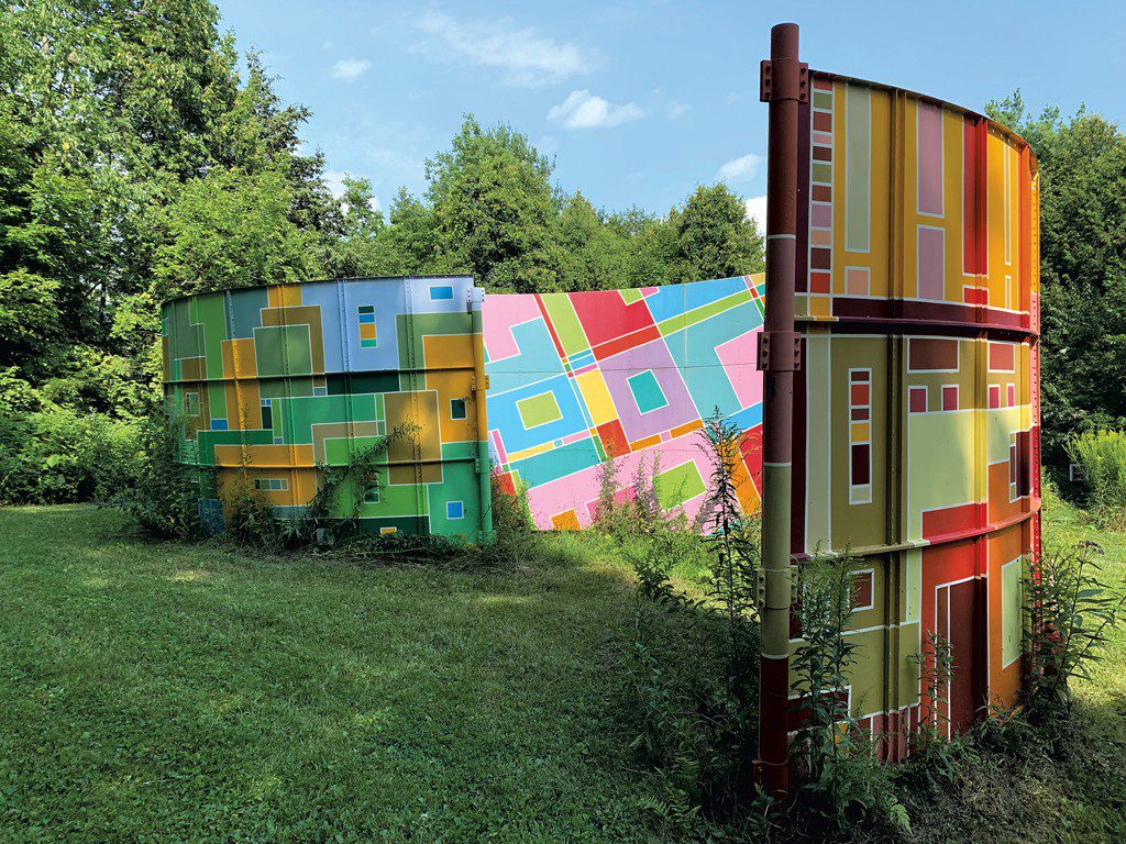 A large, curved outdoor sculpture painted with colorful abstract geometric patterns, situated in a grassy field surrounded by dense green trees under a sunny sky.