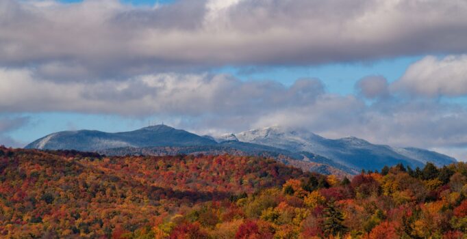 Mountains with red, orange, and yellow leaves are in the foreground of the image, while other mountain peaks visible behind have snow on them. The sky is blue with some clouds.