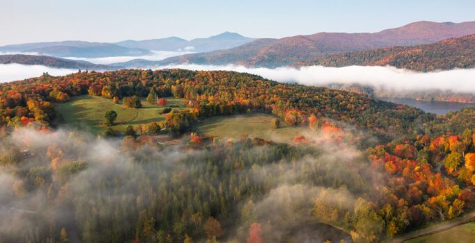 Seen from above, a landscape in autumn with mist over mountains and red, orange, and yellow leaves beginning to show up on trees.