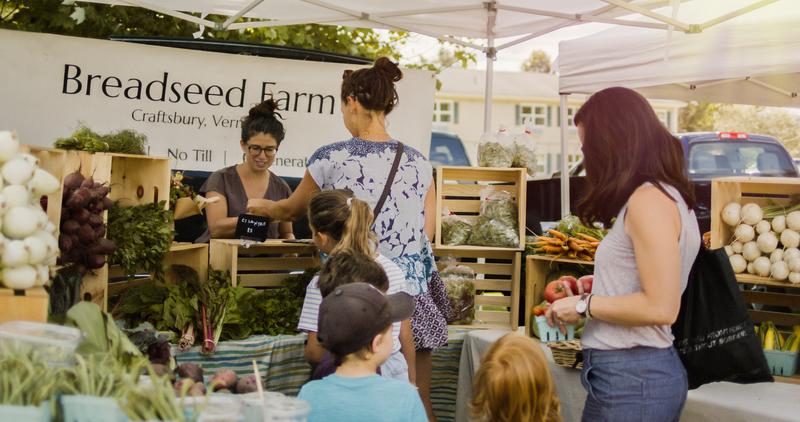 People browse inside a tent with produce and a sign reading “Breadseed Farm” at a market in the summer.