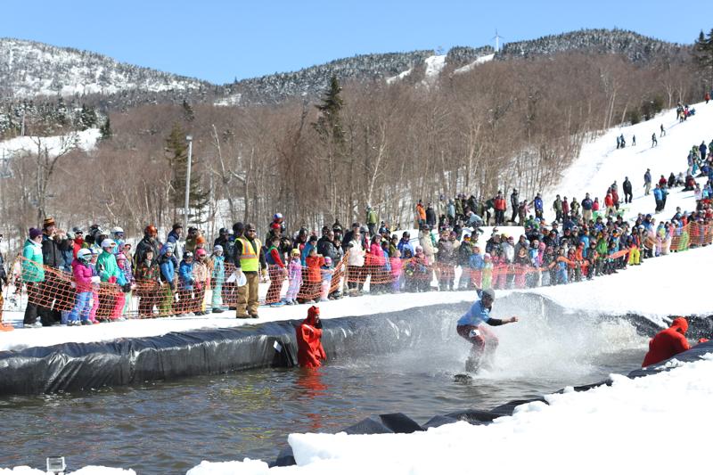 A person on a snowboard splashes down into a man-made pond at a ski resort in late winter. Crowds wearing colorful clothing celebrate.