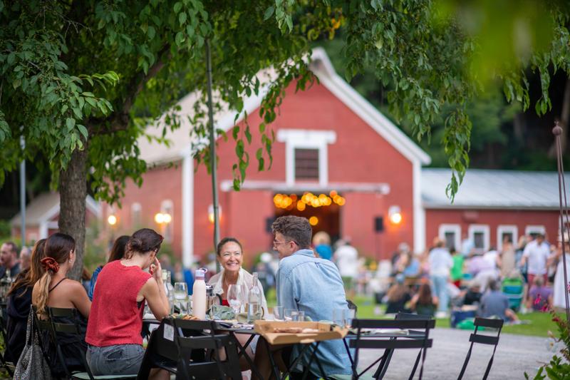 People dining at outdoor tables at The Tillerman Inn and Restaurant in Bristol, Vermont, with a historic red barn in the background.