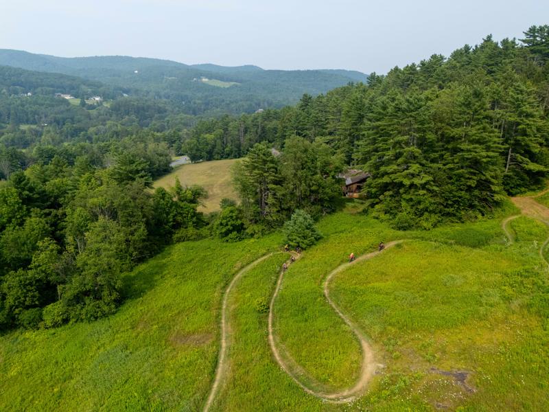 Seen from above, two people on bikes on a mowed trail through grass on a summer day.