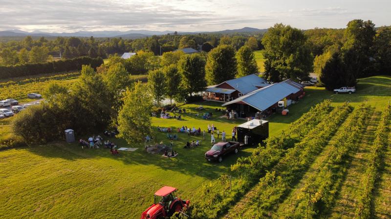 An aerial drone view of a crowd gathered on the lawn for an event at Lincoln Peak Vineyard in New Haven, Vermont, surrounded by neat rows of grapevines.