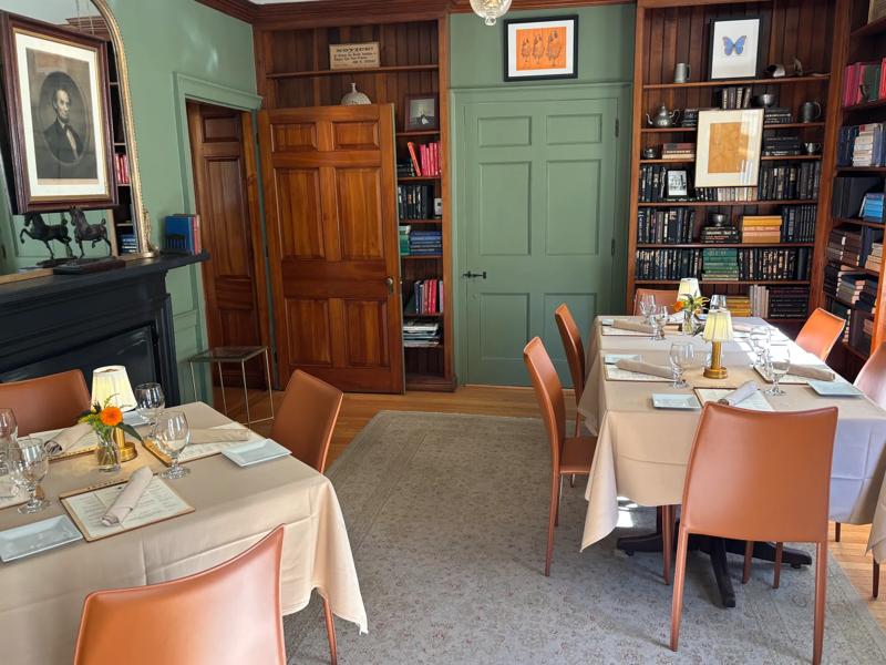 A cozy, upscale dining room, at Jessica's at the Swift House Inn, featuring set tables with orange leather chairs, sage green walls, and built-in wooden bookshelves.