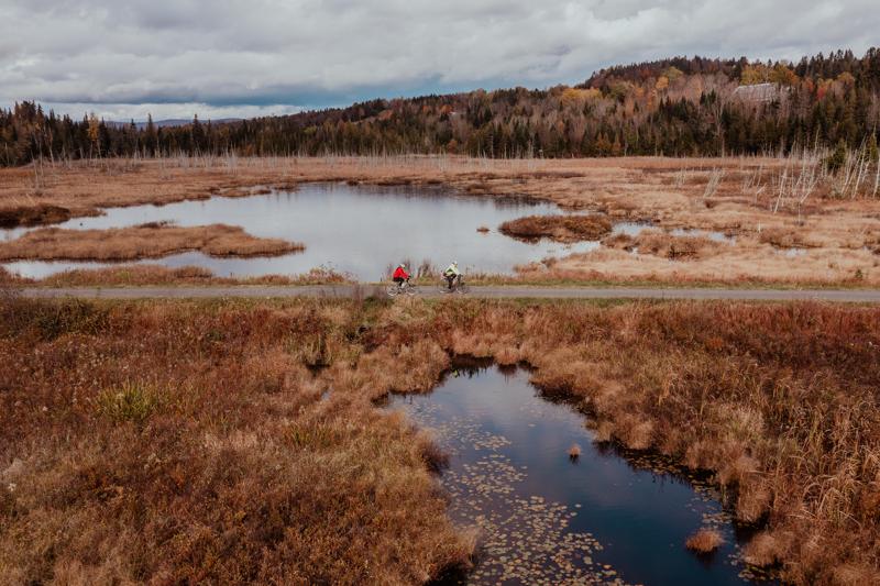 Seen from above, two people on bikes on a rail trail. Water is around them and trees have shed their leaves. A mountain is visible behind.