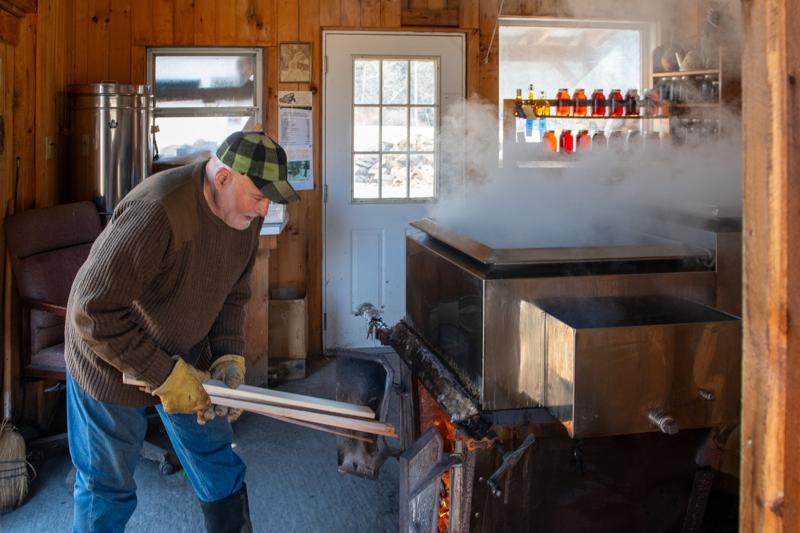 A person loads wood into a boiler in a maple sugarhouse while steam fills the room. Behind them, bottles of syrup are visible.