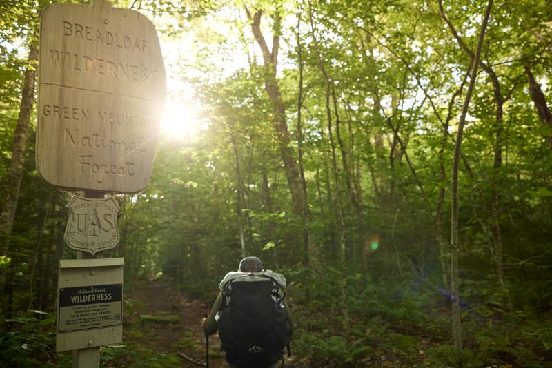 A backpacker hiking past a wooden trail sign for the Breadloaf Wilderness in Green Mountain National Forest.