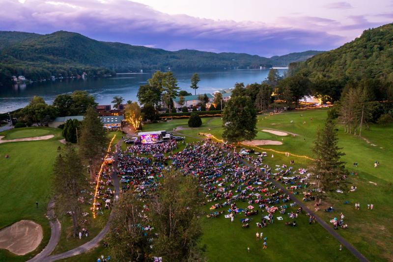 Seen from above in summer, a crowd gathers at an outdoor event by a lake. Mountains are visible on the left. A band performs at a stage.