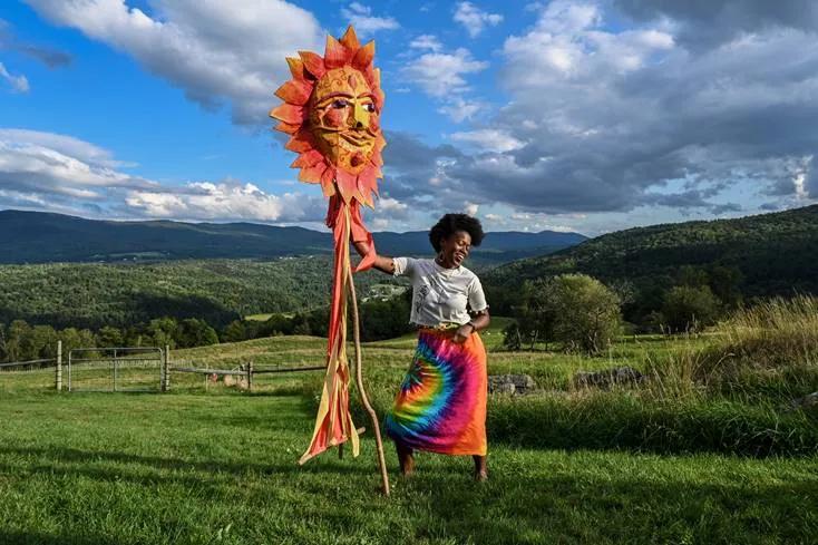 A person wearing bright clothing holds up a large puppet in a field in summer.
