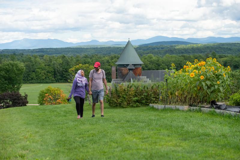 Two people walk hand in hand toward the camera through a grassy field in summer. Behind them, a building with a brick turret is visible.