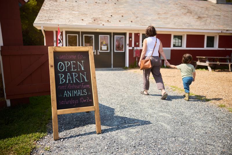 An adult and a child walk into a barn outdoors. A chalkboard sign reads “Open Barn.”