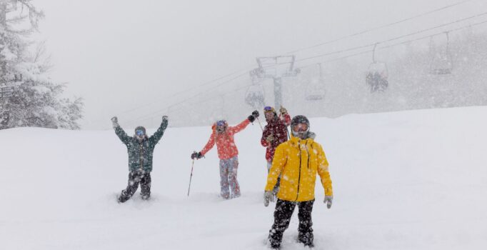 Family of four show off excitement with their hands in the air on a snowy winter day. Faintly in the background, people are transported up the ski mountain on a chairlift.
