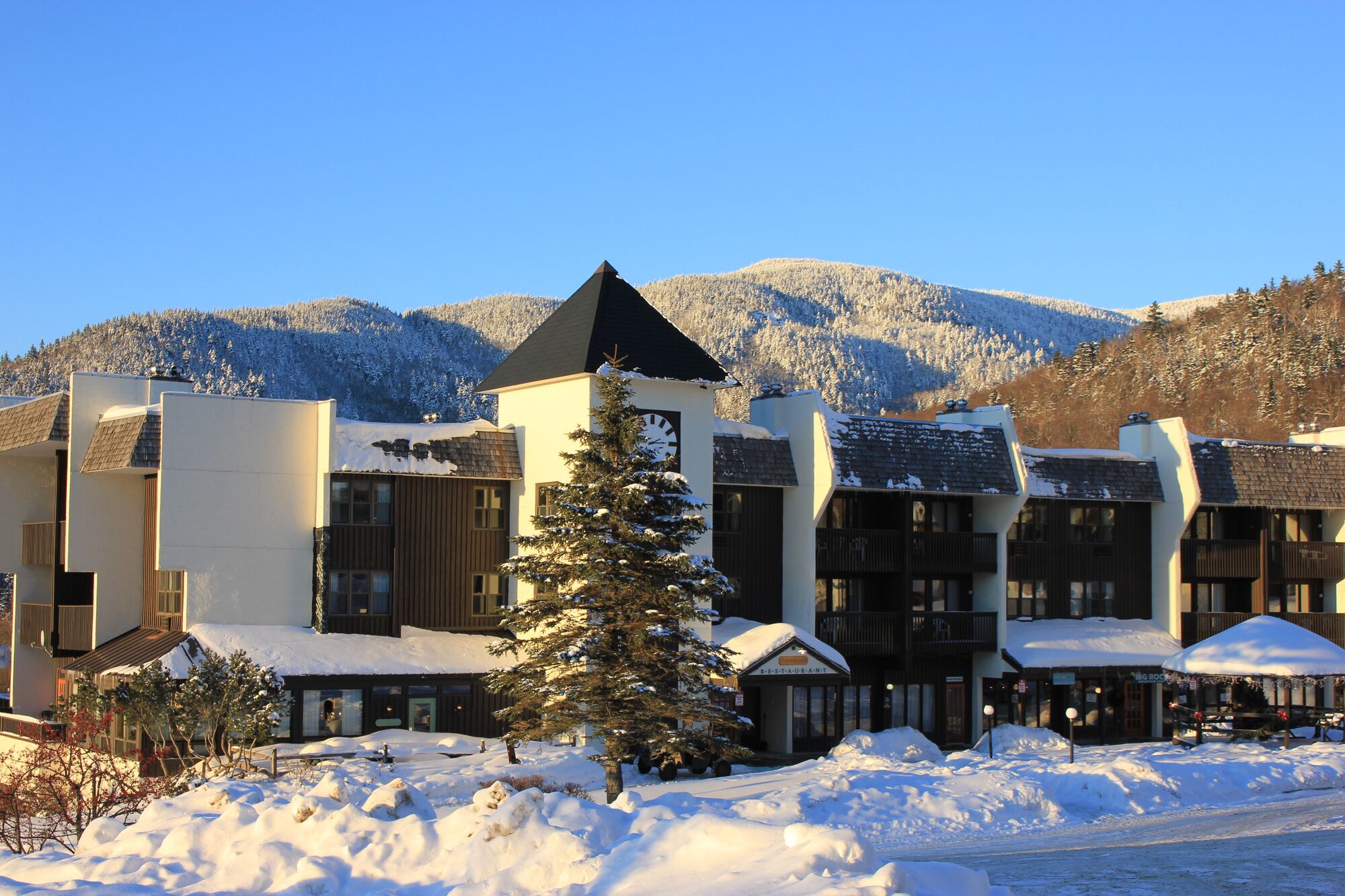 A ski lodge is glinted with sunlight. Fresh snow covers the roof and ground outside. In the background, snow-covered mountains rise up.