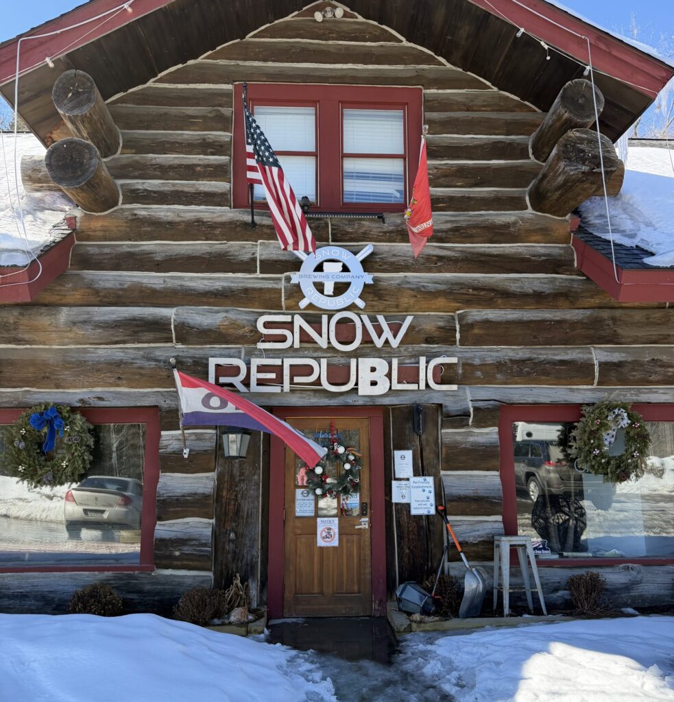 A two story log cabin has a sign out front that says “Snow Republic” as well as an “open” sign waiving in the wind.