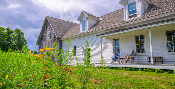 Two people sit on the porch at a white farmhouse seen from outside. Nearby, colorful flowers bloom on a sunny summer day. 