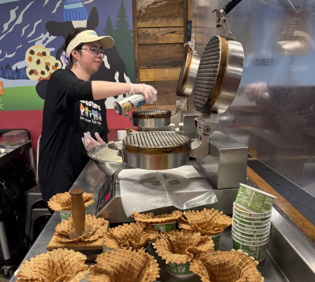 Person stands in front of two waffle making machines. Eight waffle hard shells are stuffed into cups, prepared for ice cream to be scooped into them.