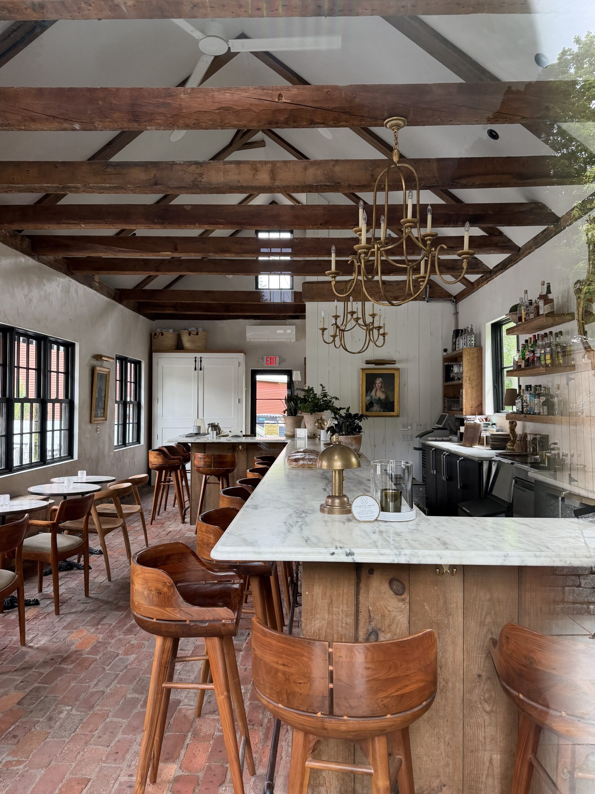 A post and beam building has brick floors and a marble bar top. Wooden stools line up to the bar, and tables are set beside them.