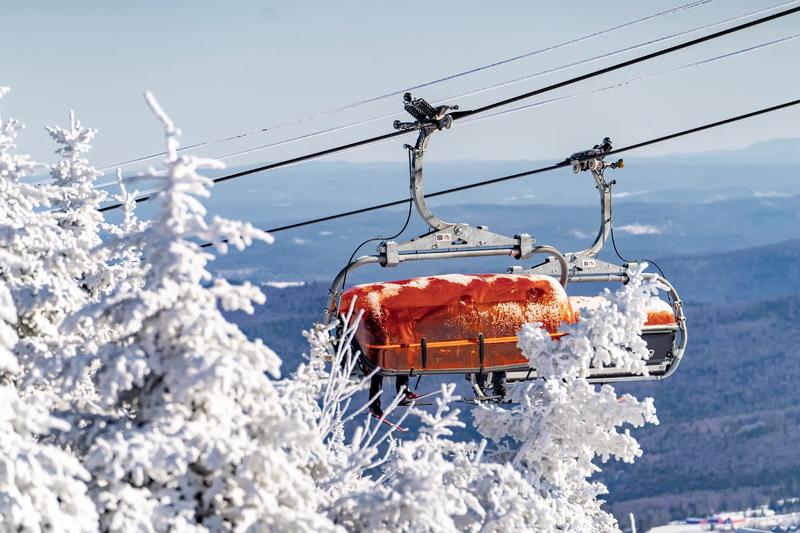Skiers riding a covered chair lift with snowy trees in the foreground.