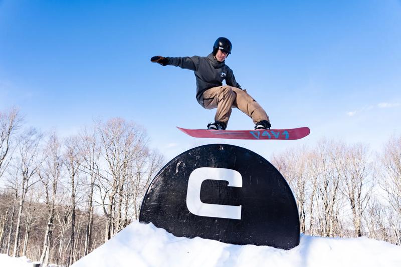 A snowboarder clears a feature in a park on a sunny day. The feature has a large C written on it. 