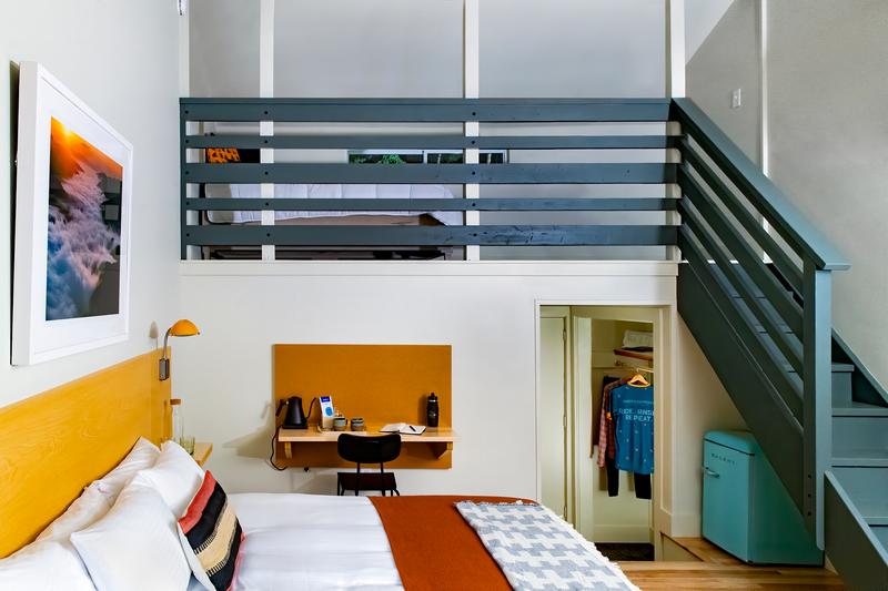 Interior of a hotel room showing a queen bed on the main floor with a desk and closet behind it and staircase going upstairs to another bed.