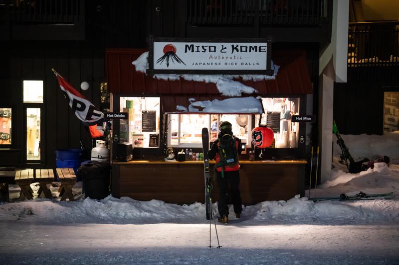 A skier orders food at a bright window outside at night. The sign says Miso Kome Authentic Japanese Rice Balls.