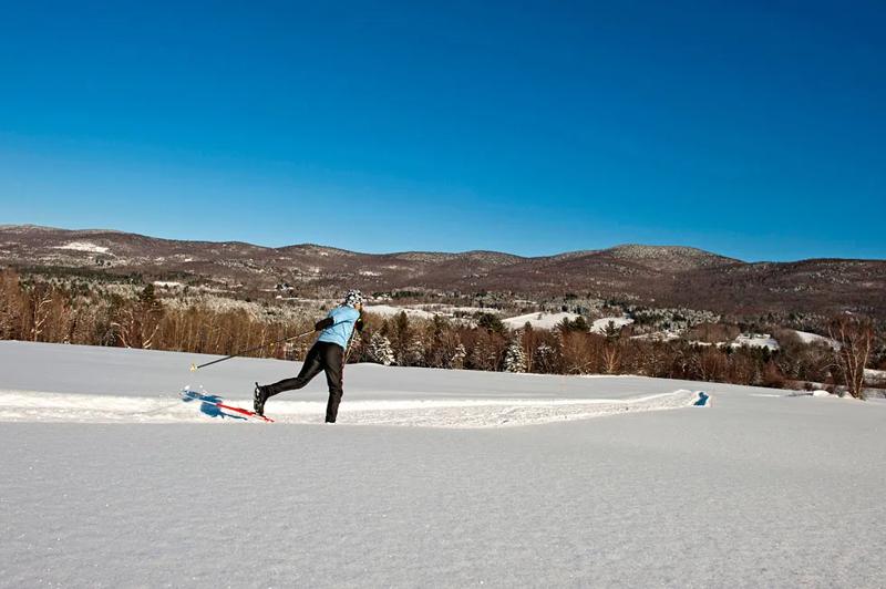 Cross country skier glides on a groomed trails. Fresh snow surrounds the trail and the sky is blue.