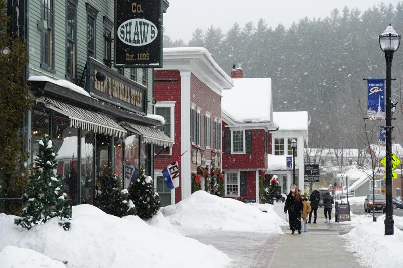 People walk on the sidewalk as open flags and wreaths hang on store windows. Snow falls and gathers on roofs. 