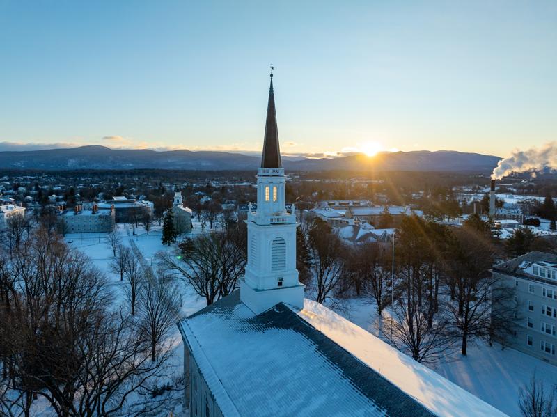 A steeple stands tall over a town on a winter day. The sun rises above the mountains in the distance.
