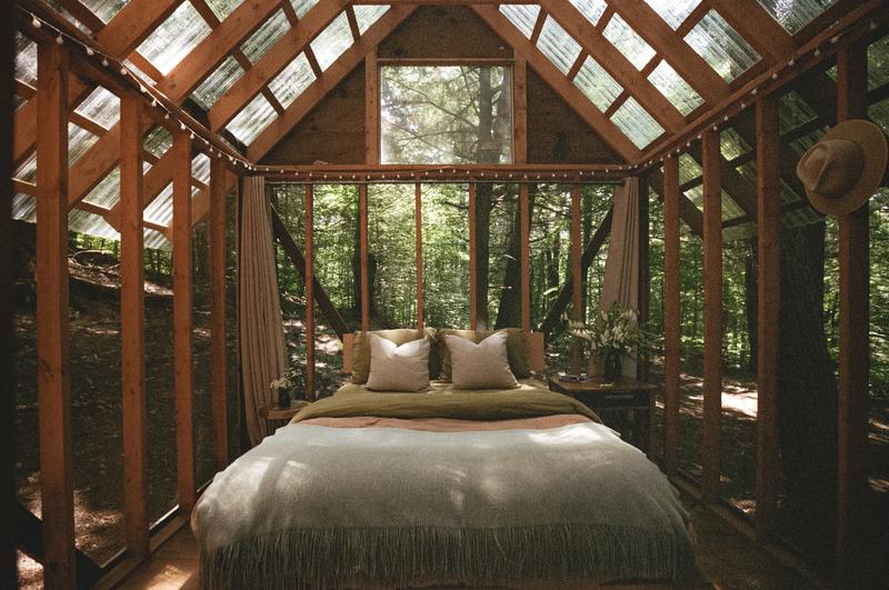 A large bed with neutral-toned bedding inside a greenhouse in summer.